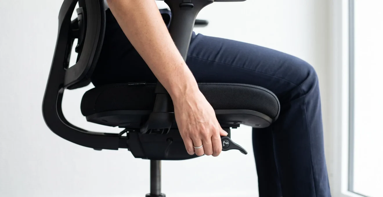 Gros plan sur main de femme actionnant le levier de réglage de profondeur d'assise situé sous l'assise noire d'un siège ergonomique, vue de côté avec jambes partiellement visibles